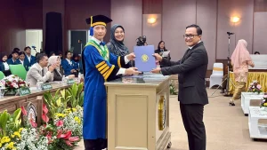 The Dean of the Faculty of Medicine, wearing a blue academic gown, hands over a blue diploma folder to a male graduate in a black suit.