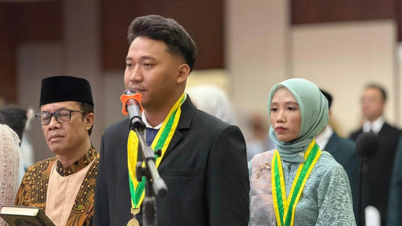 A close-up of a male graduate reciting the professional oath into a microphone, accompanied by a religious official and a female graduate standing beside him.